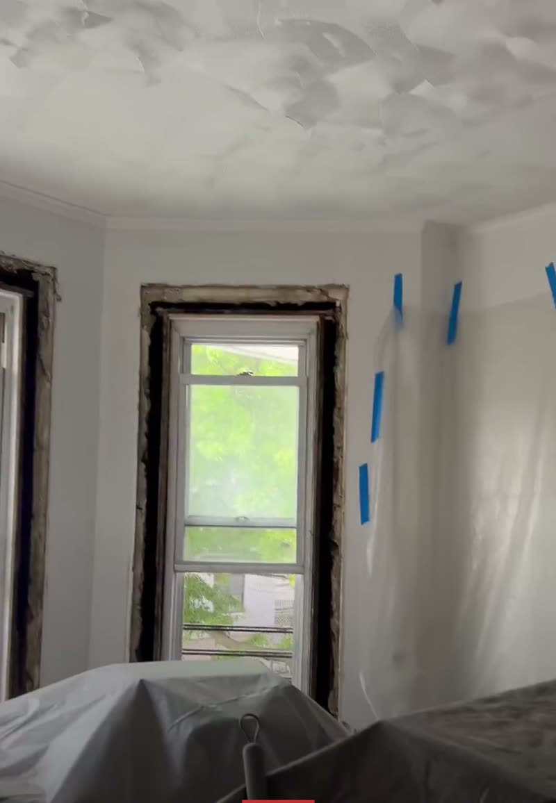 Before: apartment room with the window casing stripped off, patchy ceiling plaster and furniture covered in plastic sheeting